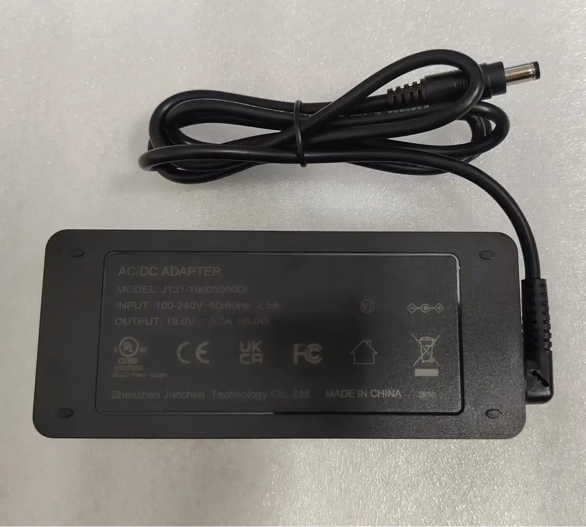 A black DC power adapter with its attached cable, photographed from above on a light gray textured surface.