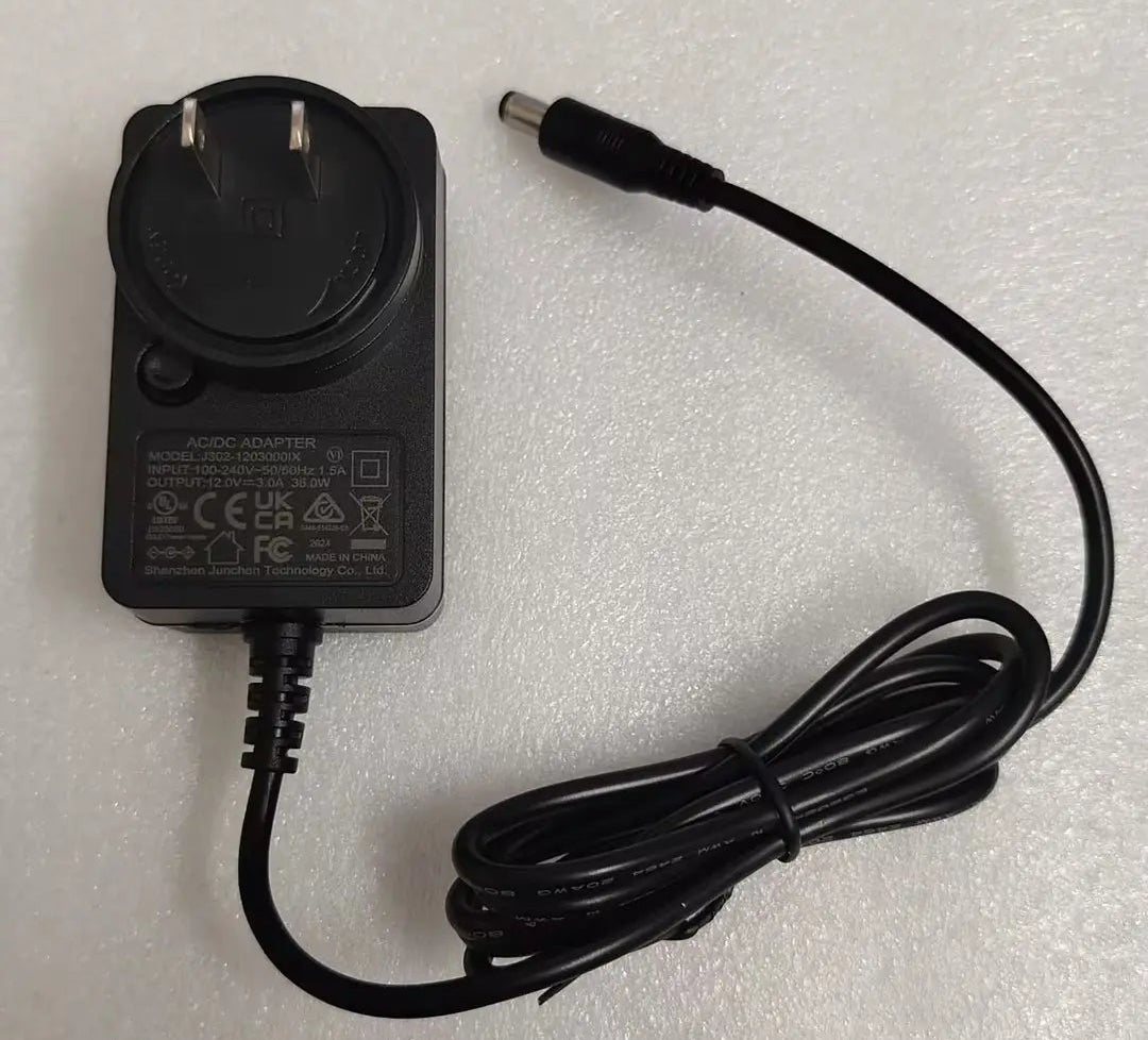 A black AC adapter power supply with a UK two-pin plug and a coiled black cable, photographed on a light gray textured surface.