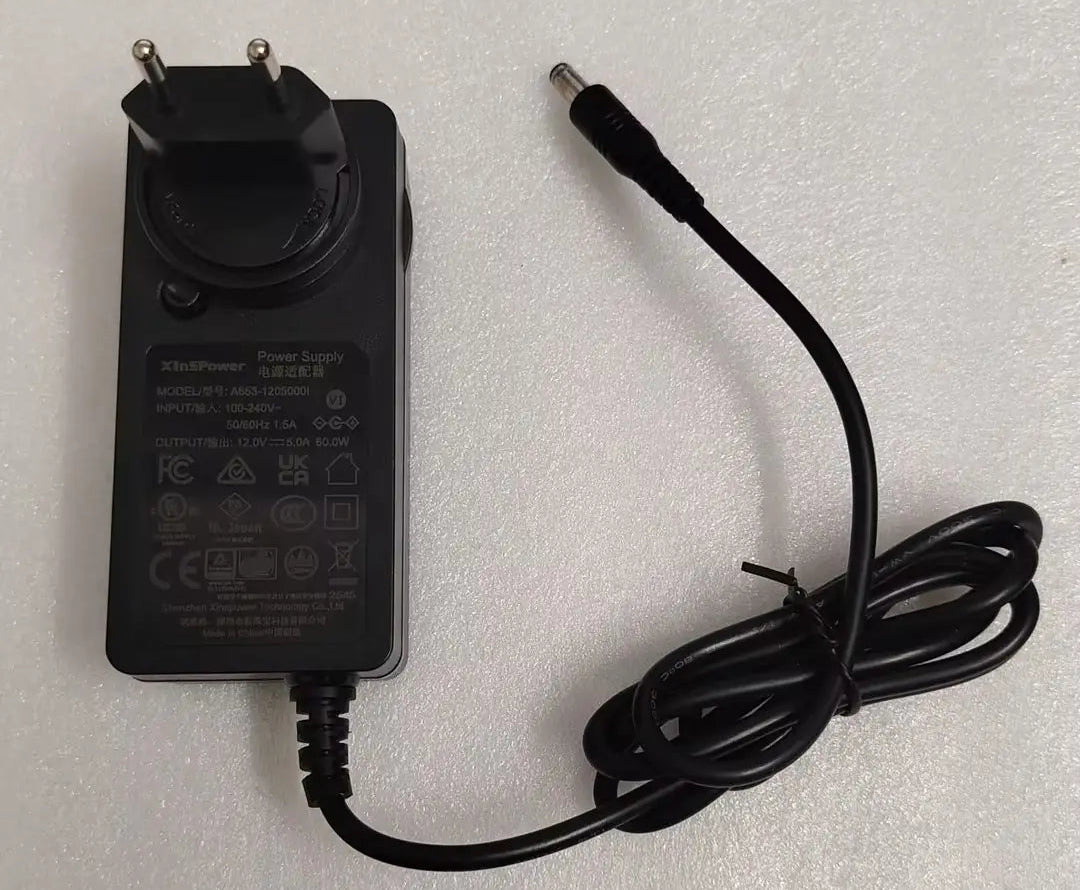 A black power adapter with a two-pin plug and a coiled black cable, photographed on a light gray textured background.
