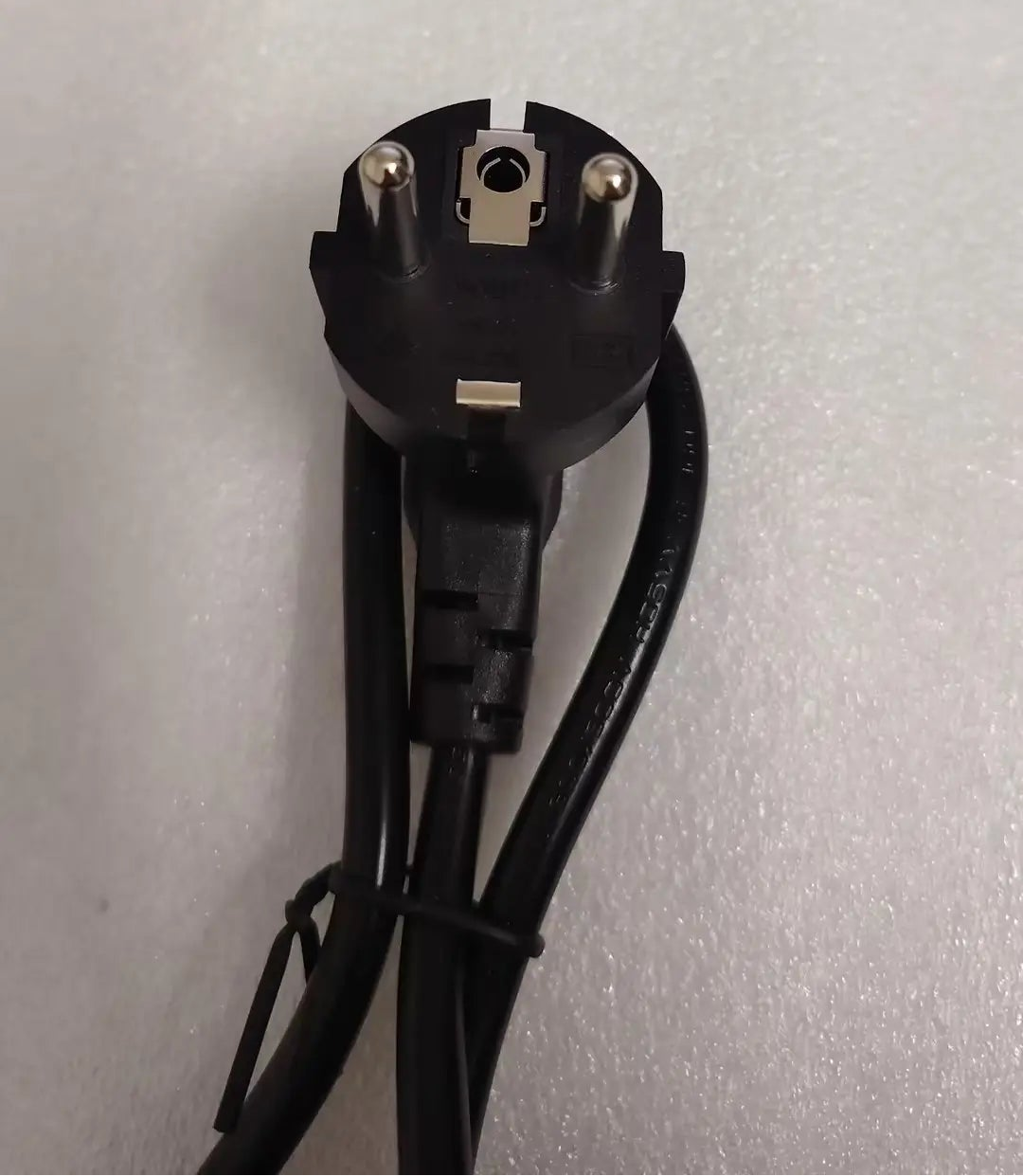 A black three-pin electrical power plug with attached black cords, photographed on a light gray textured surface.
