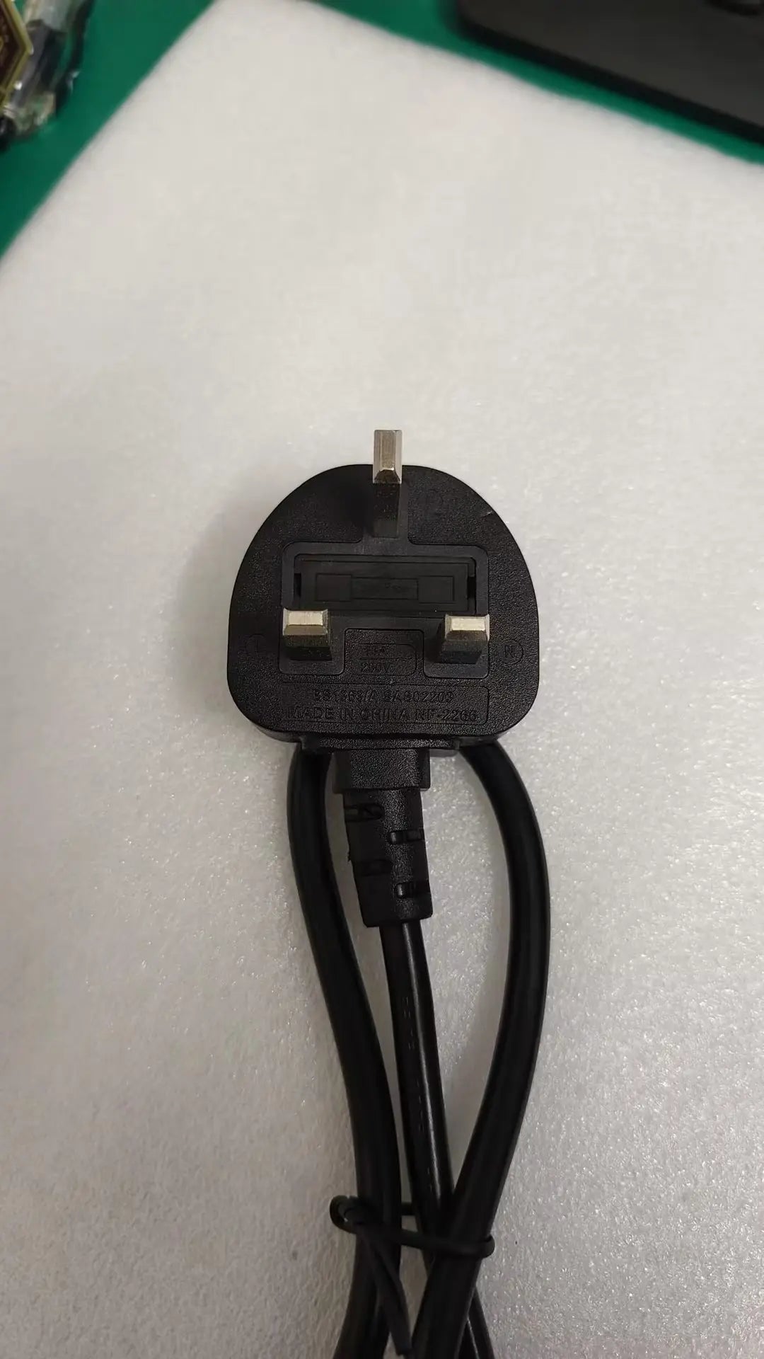 A black UK three-pin wall plug power adapter with attached black cables, photographed on a white textured surface.